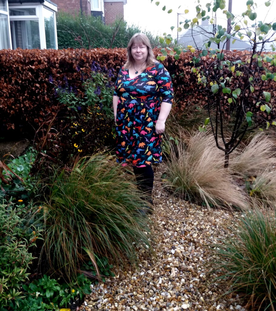 A white woman with blonde hair standing on the gravel path of her front garden. You can see grasses, perennials and a Quince tree, with a beech hedge behind her.