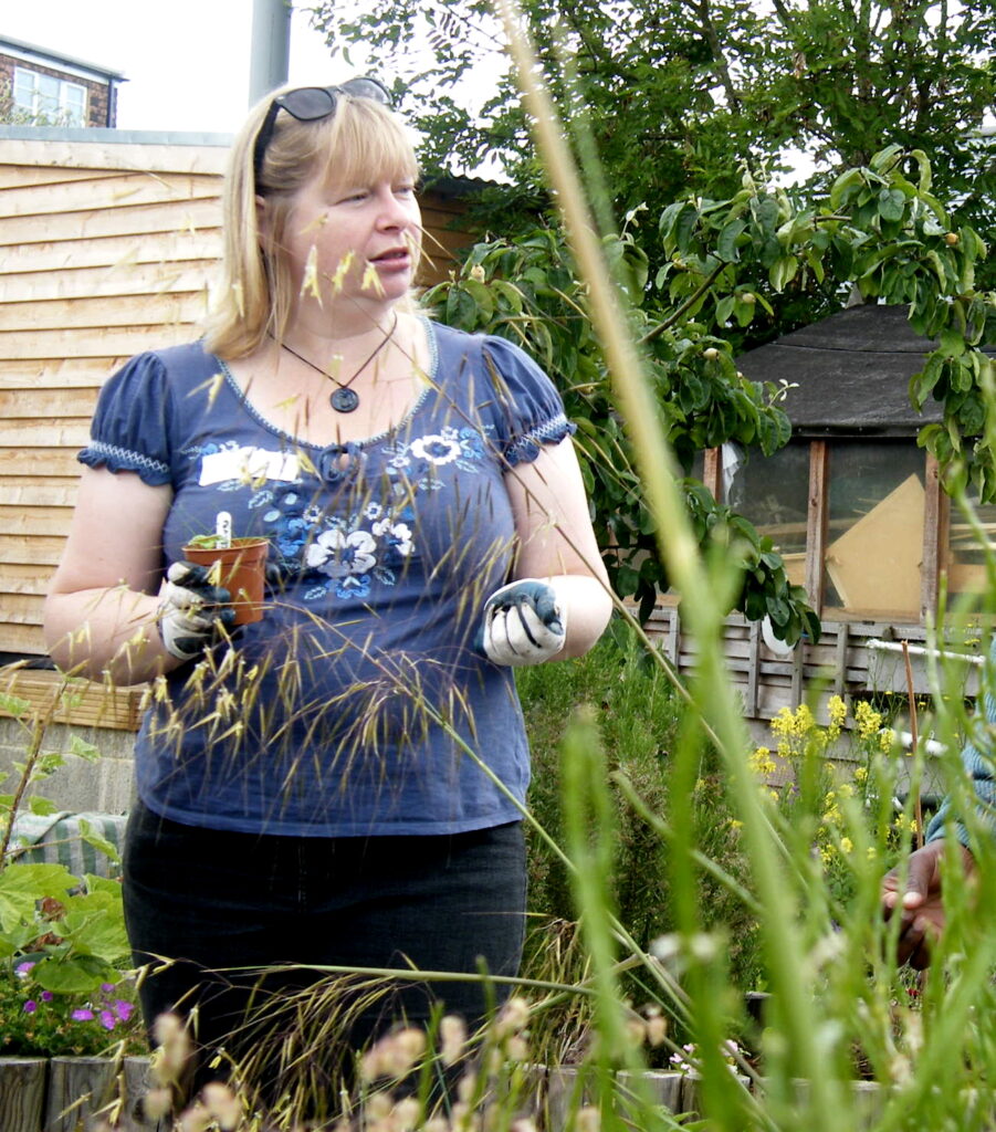 A white blonde woman standing in the community garden, wearing gardening gloves and holding a small pot with a young plant in it. She is looking to the right, and is talking about potting on or planting out young plants.