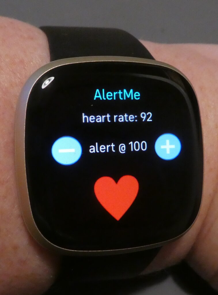 This is the AlertMe app on my Fitbit. Again it has a black background screen, with AlertMe in turquoise, then on the next line: heart rate: 92 in white. In the middle in white is: alert @ 100 with a minus and plus sign on each side and under this a red heart shape.