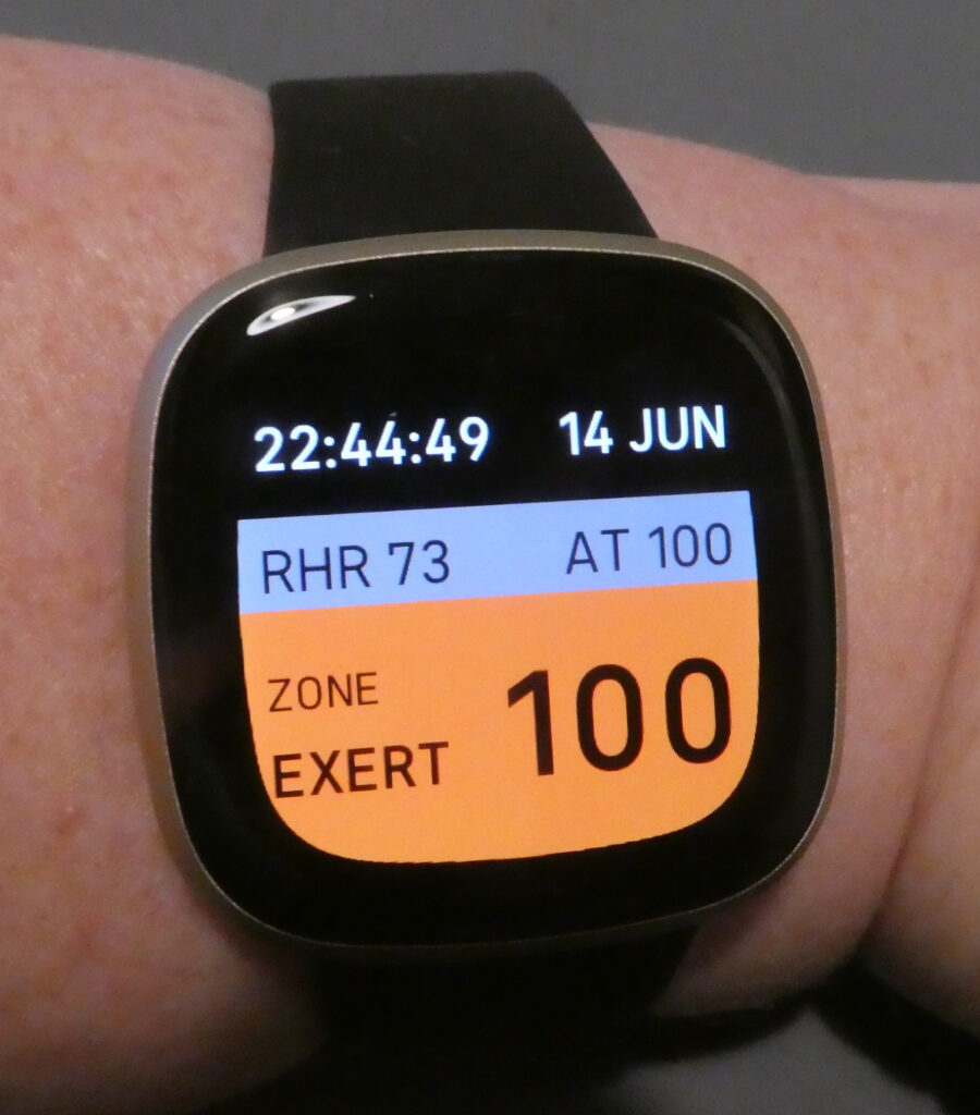 A large watch screen on a white wrist. It has a black background, with the time and date in white at the top. In the middle, in a grey panel it has the RHR, resting heart rate, and the AT 100, the bpm threshold I set. Then there is an orange panel that says Zone Exert on the left and the number 100 on the right. I've just reached my anaerobic threshold.