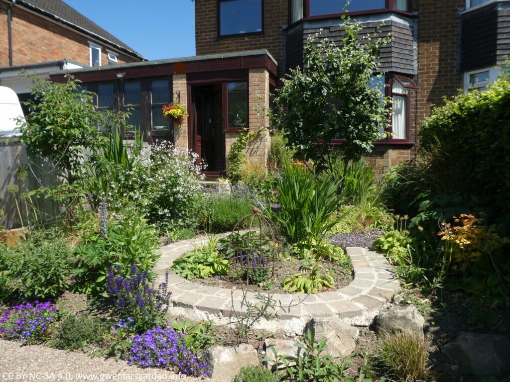 An overview of part of the front garden from the right side of the treet. There is a mix of planting including two young trees, a horse-shoe shaped paved path, more plants and the house to the rear.