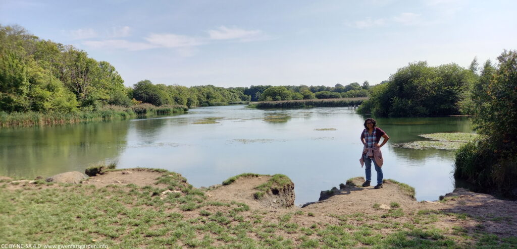 An overview of one of the lakes in September, with all the foliage on the trees surrounding it. Standing near the edge is a brown person with hands resting on their hips and a big smile. The sky is a light blue with soft clouds.