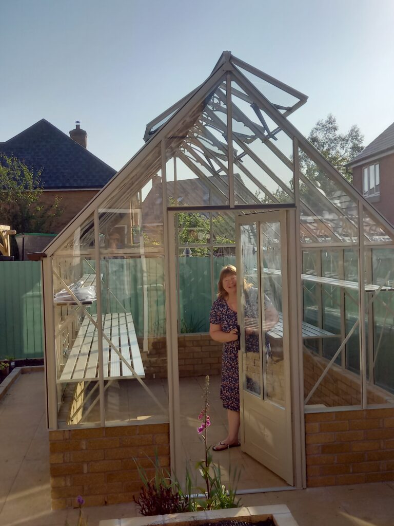 A white person in a light sage green, greenhouse, that sits on a sandy coloued bricked wall. The person is smiling and very happy.