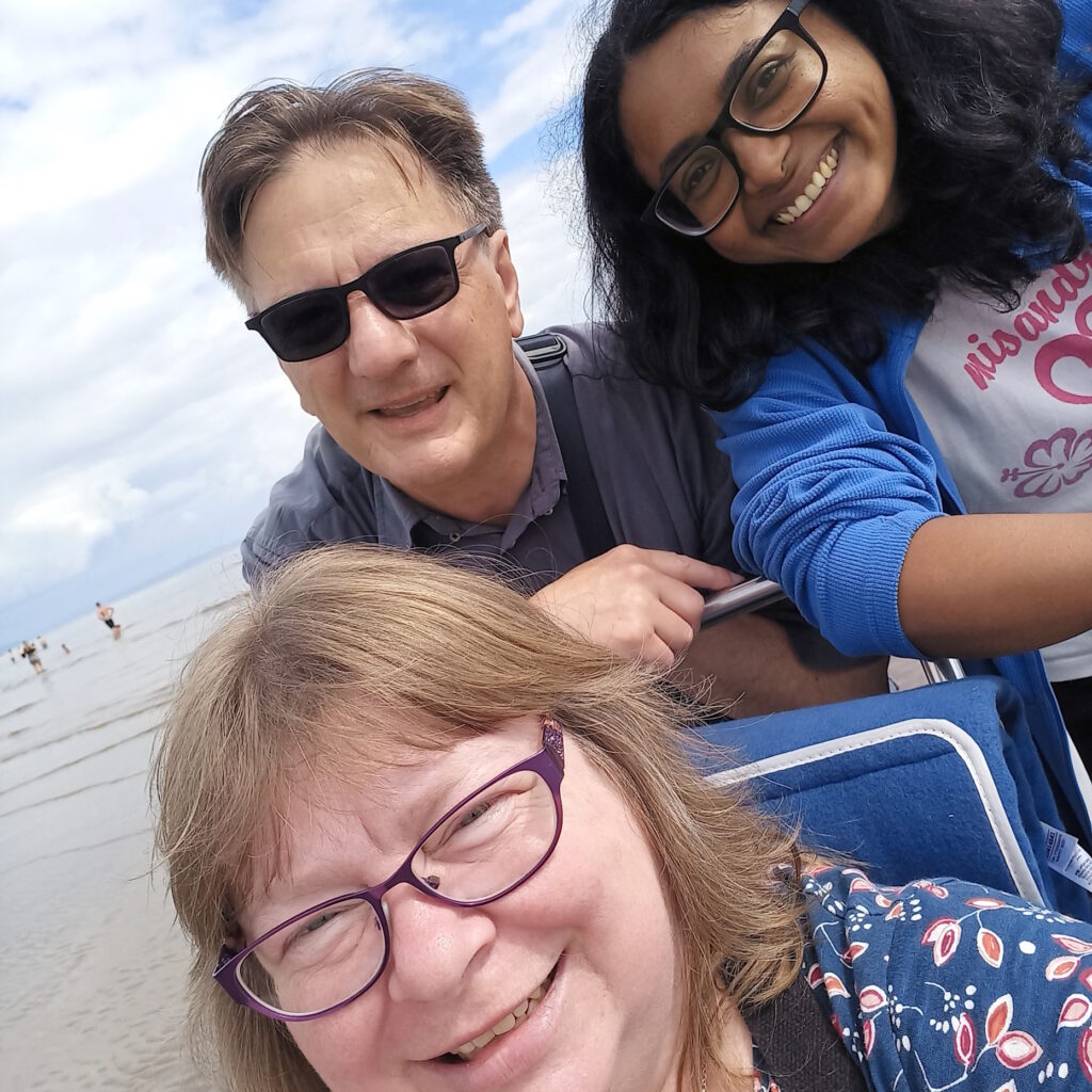 Three people, two white, one a person of colour. One white person sitting in a beach wheelchair, the other two people above and behind. Everyone is smiling and happy. To the left you can see the sandy beach and the edge of the water.