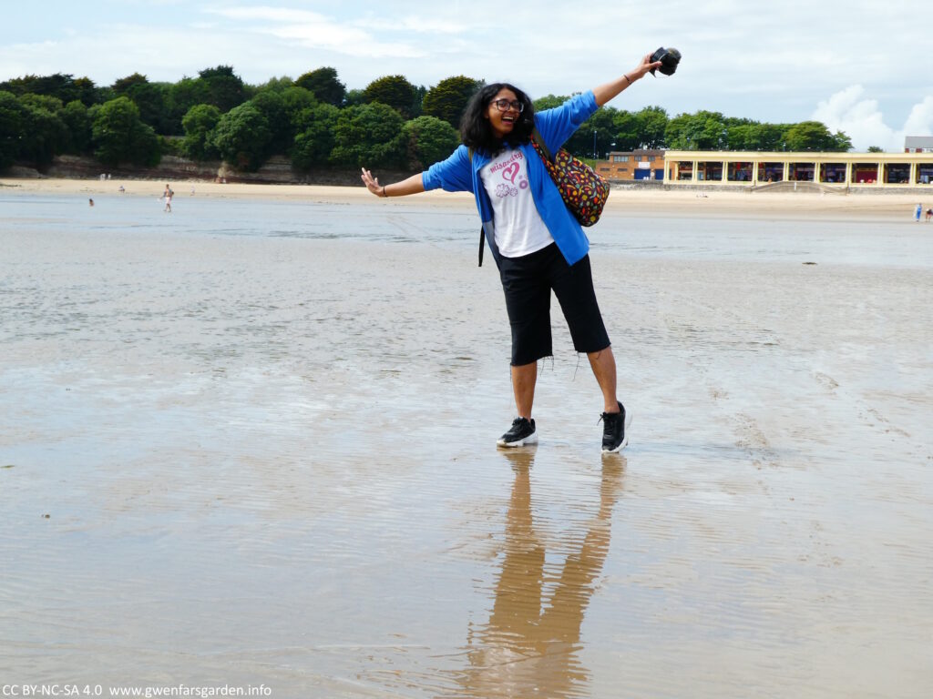 A person of colour standing on wet sand with their arms out and a big smile.