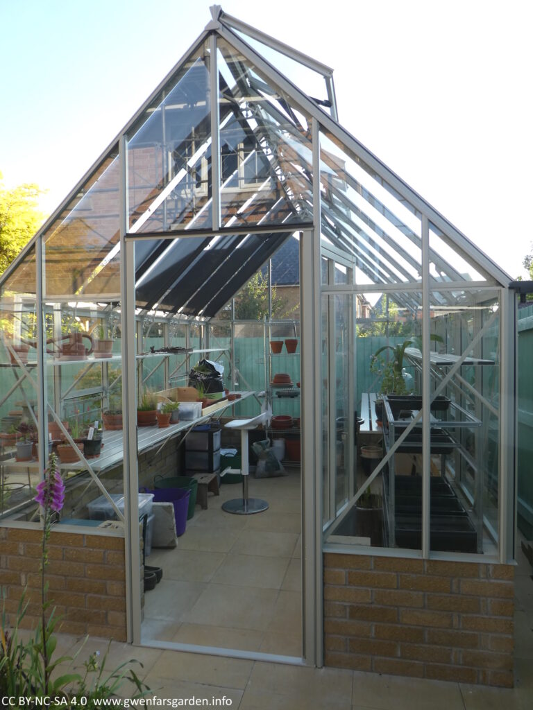 A view from the front of the greenhouse with the door open. Inside you can seen various plant related tools and a stool to sit on when potting up plants.