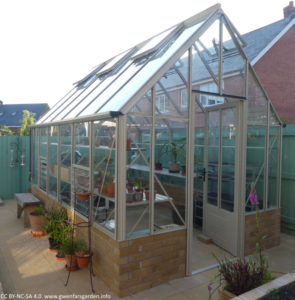 A view of the whole greenhouse looking from the front left to the back left side. You can see some plants are already inside the greenhouse. There are some plants and a wooden seat alongside the outside of the greenhouse wall.The greenhouse is made of strengthened aluminum and sits on a small (c. 80cms high) brick wall. The frame is a soft pale sage colour. 