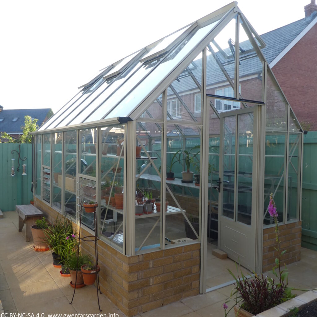 A view of the whole greenhouse looking from the front left to the back left side. There are some plants and a wooden seat alongside the greenhouse wall.