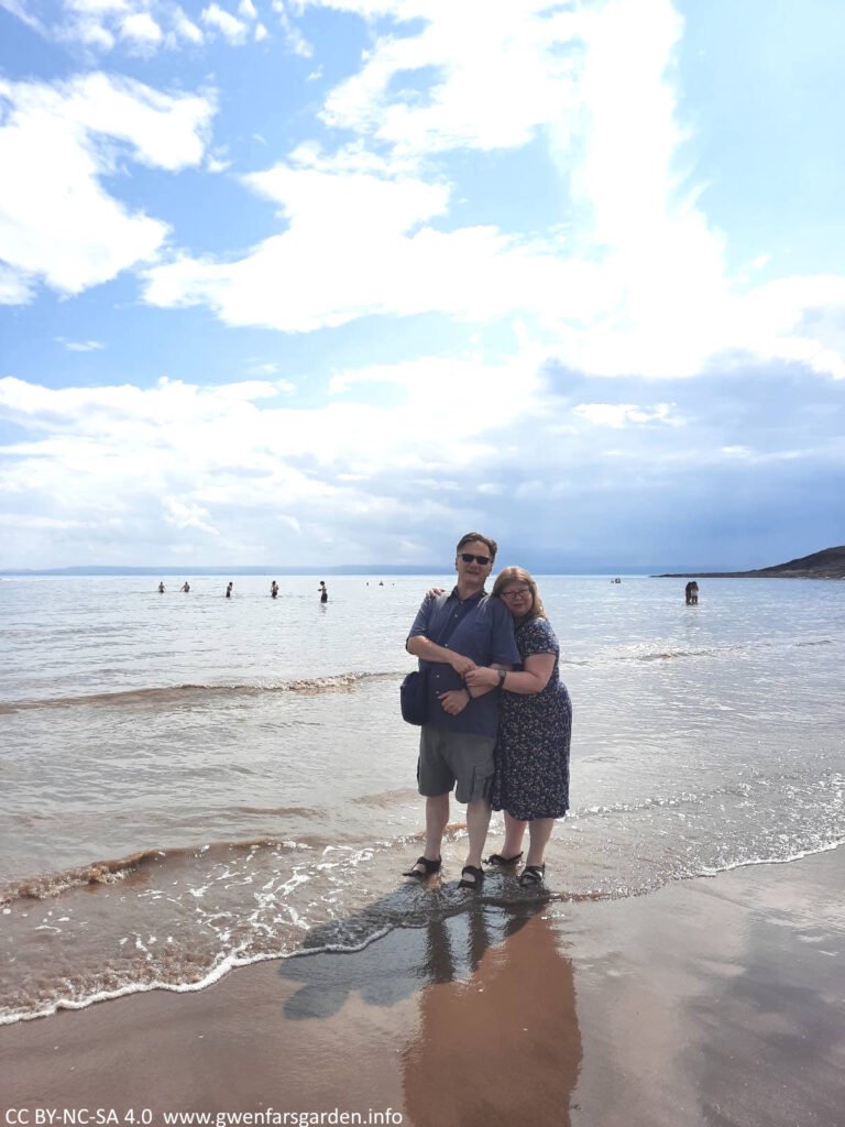 Two white people standing just inside the water line, one slightly behind and hugging the one in front. Both are smiling and are happy.