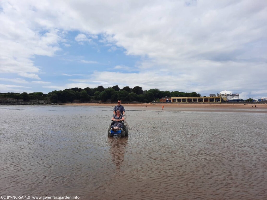 Two white people on wet sand. One is standing and pushing the other in the beach wheelchair.