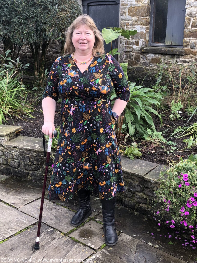 A white person with a walking stick standing in a garden.