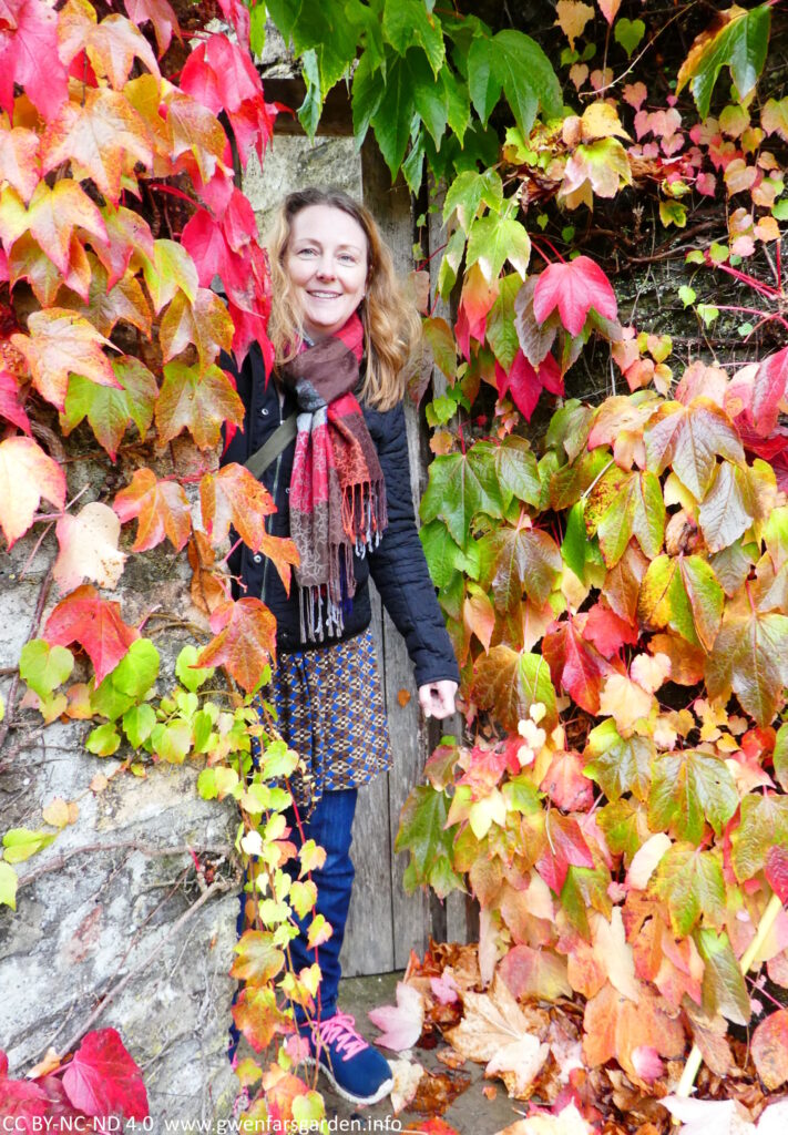 A doorway with lots of Virginia Creeper foliage. A white person is partially stepping through and smiling at the camera.