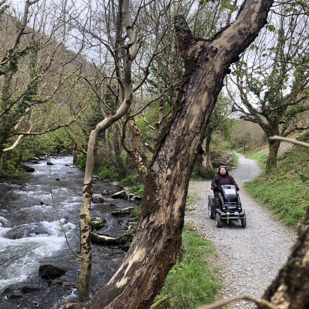 A smiling white person on a tramper mobility scooter, along a stony path, surrounded by lots of fresh green spring growth of ferns and young tree leaves. There is a fast moving stream with trees along it's shore, to the left.